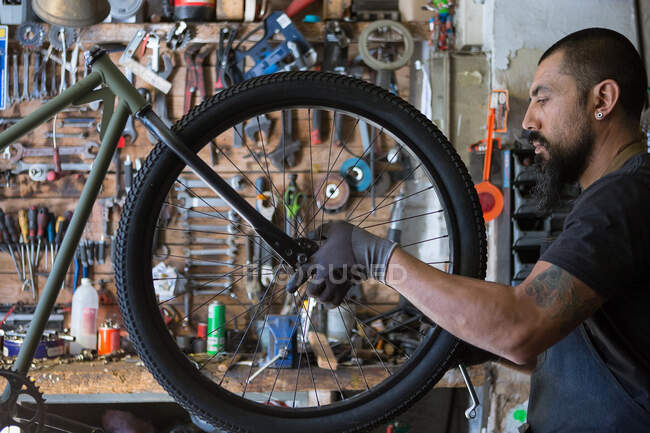 Hombre adulto serio en delantal y guantes reparando rueda de bicicleta en garaje moderno - foto de stock