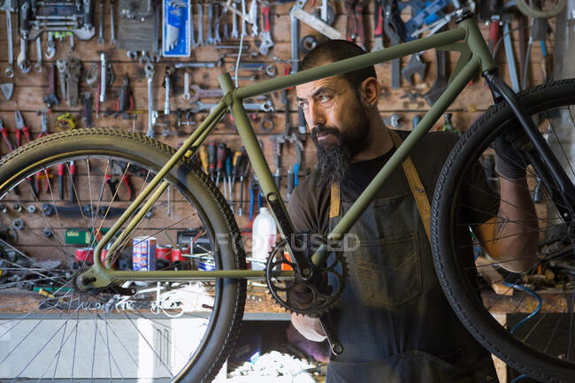 Hombre adulto serio en delantal y guantes reparando rueda de bicicleta en garaje moderno - foto de stock