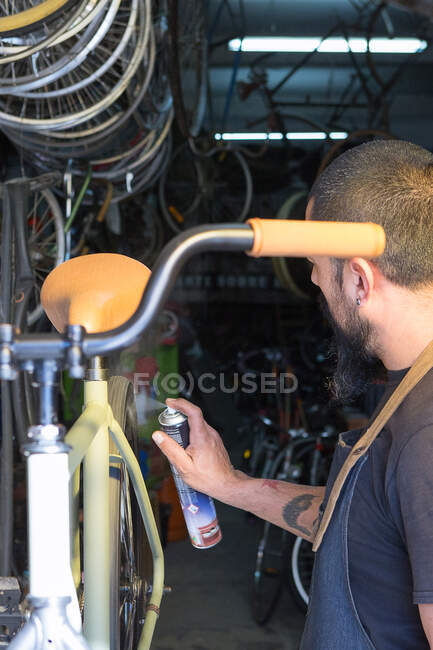 Homme barbu dans le tablier avec l'huile de pulvérisation d'équipement sur la roue tout en réparant le vélo dans le garage — Photo de stock
