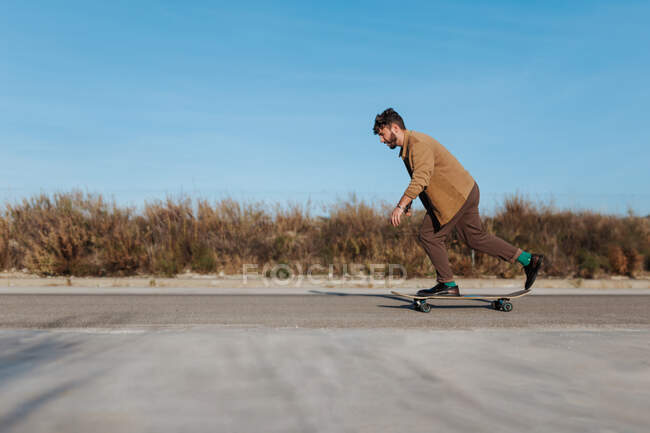 Вид збоку на повне тіло молодий бородатий чоловічий фігурист у стильному одязі їзда скейтборд уздовж асфальтової дороги в сільській місцевості — стокове фото