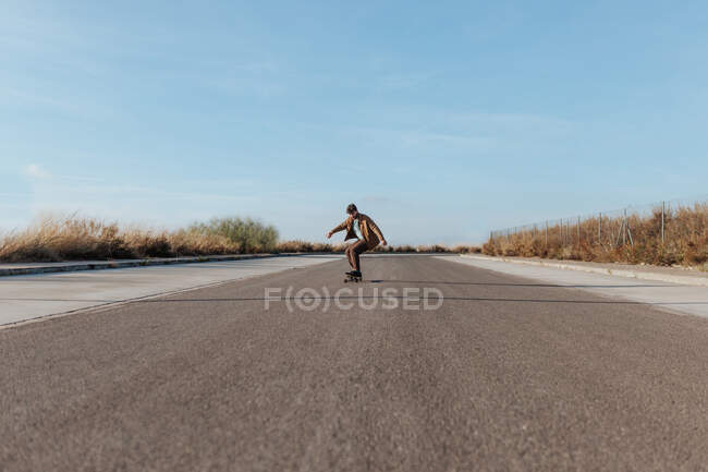 Full body young bearded male skater in stylish wear riding skateboard along asphalt road in countryside - foto de stock