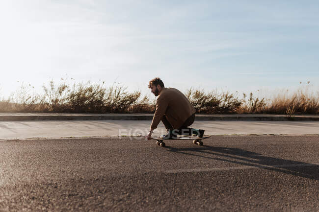 Молодий бородатий чоловічий фігурист у повсякденному одязі виконує трюк, торкаючись землі під час їзди на асфальтній дорозі — стокове фото