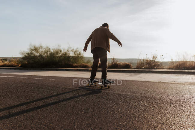 Full body back view of anonymous male skater in stylish wear riding skateboard along asphalt road in countryside — Stock Photo