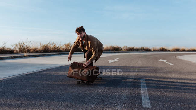 Full body serious young bearded male skater in trendy clothes with leather bag riding skateboard along asphalt road — Foto stock