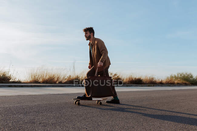 Side view of full body serious young bearded male skater in trendy clothes with leather bag riding skateboard along asphalt road - foto de stock