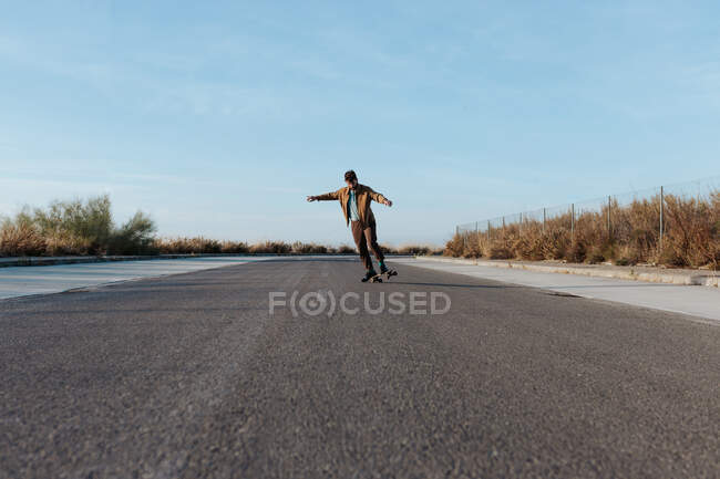 Full body young bearded male skater in stylish wear riding skateboard along asphalt road in countryside — Foto stock