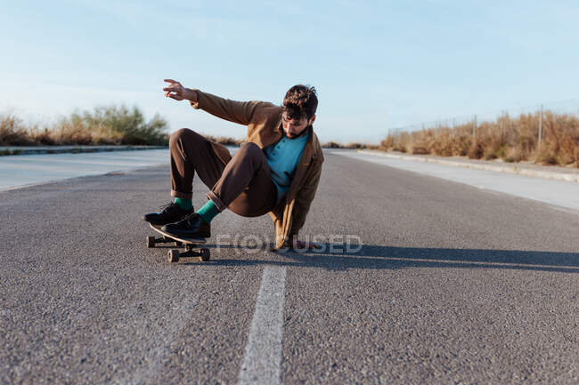 Full body young bearded male skater in casual clothes performing trick touching ground while riding on asphalt road - foto de stock