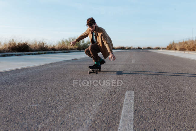 Full body young bearded male skater in casual clothes performing trick touching ground while riding on asphalt road — Foto stock