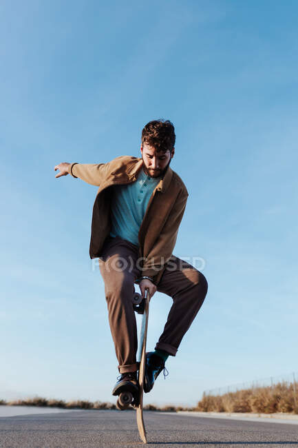 Full body young bearded male skater standing on edge of skateboard keeping balance while performing trick on asphalt road with hand raised and looking down — Fotografia de Stock