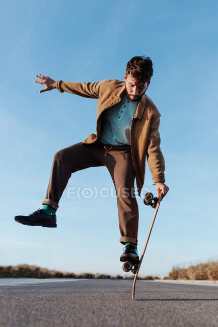 Full body young bearded male skater standing on edge of skateboard keeping balance while performing trick on asphalt road with hand raised and looking down — Foto stock