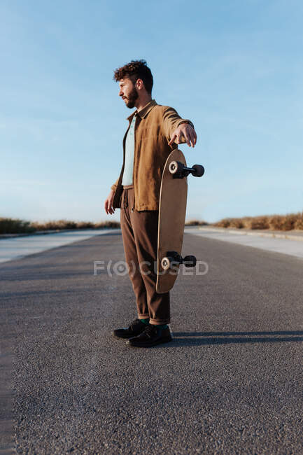 Серйозний повний бородатий чоловічий фігурист у повсякденному вбранні кидає дошку, стоячи на асфальтній дорозі і дивлячись вбік — стокове фото