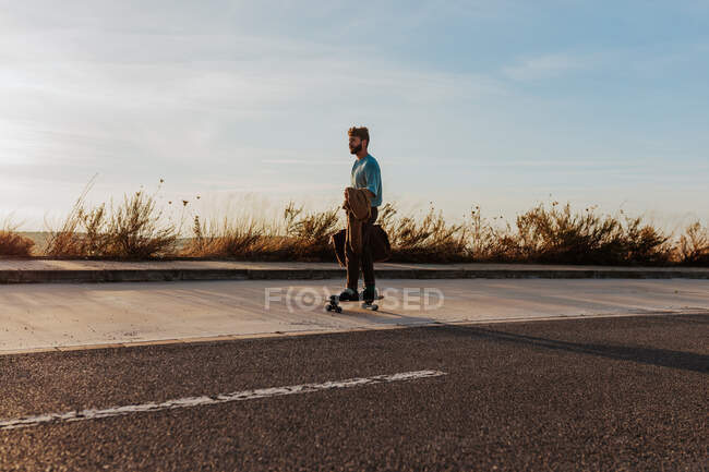 Full body young bearded male riding skateboard with bag and jacket in hand along pavement near asphalt road - foto de stock