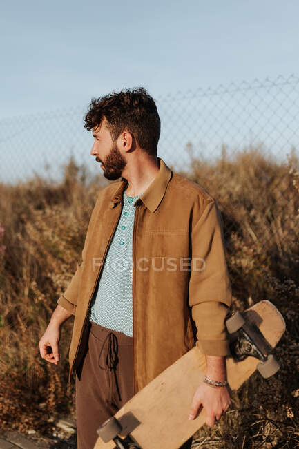 Молодий бородатий чоловічий фігурист у повсякденному одязі, що стоїть біля трави та паркану з скейтбордом, дивлячись далеко — стокове фото