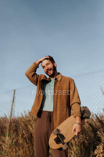 Jeune patineur masculin barbu souriant en tenue décontractée debout près de l'herbe et de la clôture avec planche à roulettes et cheveux touchants regardant loin — Photo de stock