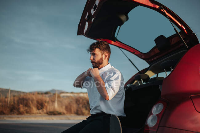 Side view of young bearded male driver sitting in opened car trunk and taking white shirt off looking away — Stock Photo