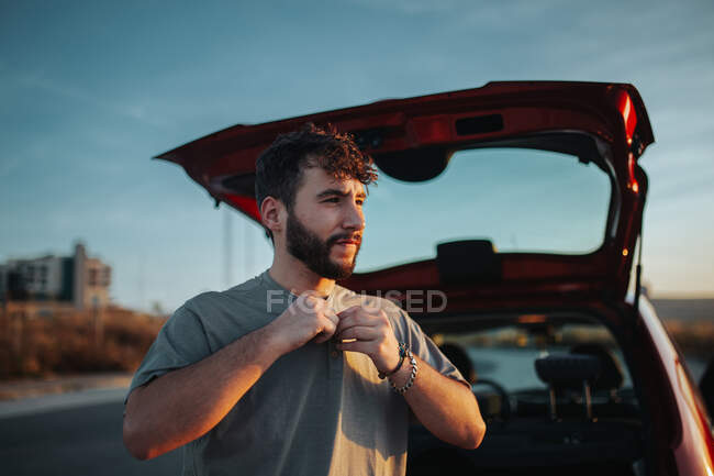 Young bearded male driver sitting in opened car trunk and putting shirt on looking away - foto de stock
