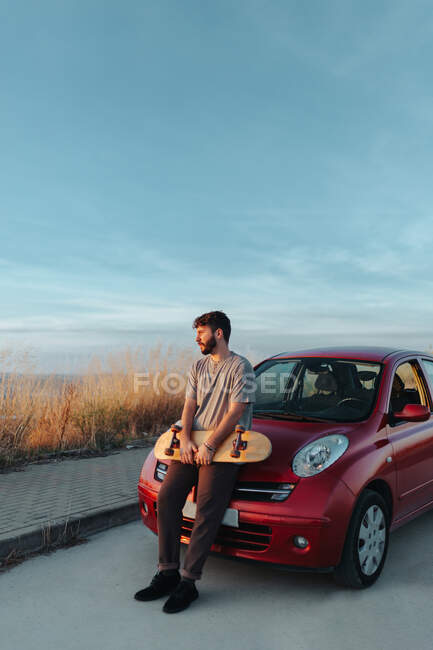 Full body thoughtful young bearded male skater in casual clothes leaning on hood of car parked on roadside with skateboard in hands and looking away - foto de stock