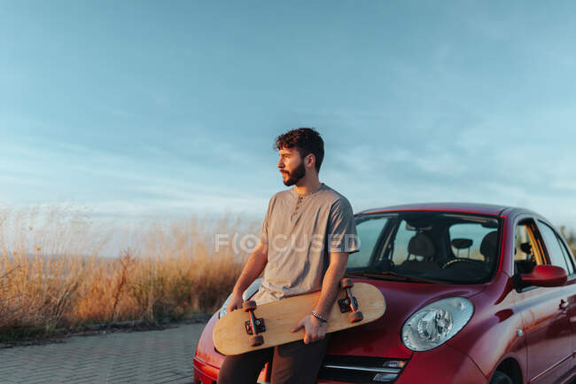 Вдумчивый молодой бородатый мужчина-фигурист в повседневной одежде опирается на капот припаркованного на обочине автомобиля со скейтбордом в руках и отворачивается — стоковое фото