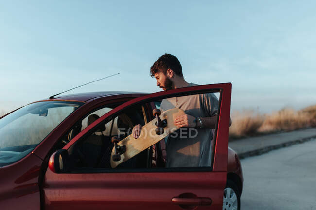 Концентрированный молодой бородатый мужчина в повседневной одежде кладет скейтборд в автомобиль, припаркованный на асфальтированной дороге в природе — стоковое фото