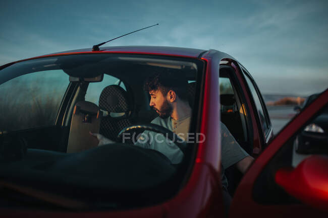 Концентрированный молодой бородатый мужчина в повседневной одежде кладет скейтборд в автомобиль, припаркованный на асфальтированной дороге в природе — стоковое фото