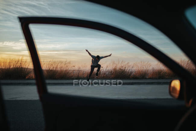 From car view of full body young man in casual wear jumping on skateboard while performing kickflip on asphalt road against dusky sky — Foto stock