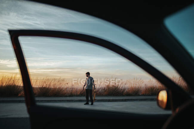 С автомобиля вид на все тело молодой человек в повседневной одежде со скейтбордом на асфальтированной дороге против сумерки — стоковое фото