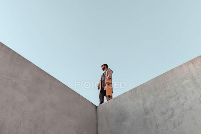 From below of young bearded male skater standing on high concrete ramp in skatepark with skateboard looking away — Stock Photo