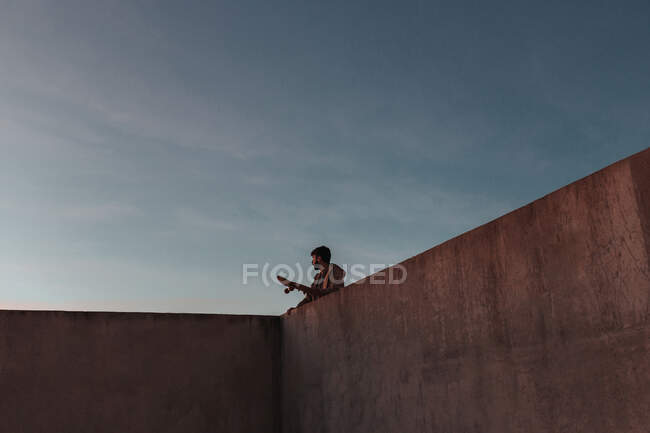 From below of young bearded male skater standing on high concrete ramp in skatepark with skateboard looking away - foto de stock