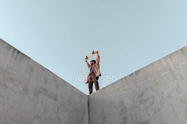 From below of young bearded male skater standing on high concrete ramp in skatepark with skateboard above head and looking away — Stock Photo