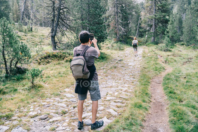 Back view of unrecognizable male backpacker taking photo of female partner on cellphone on pathway between green trees — Stock Photo