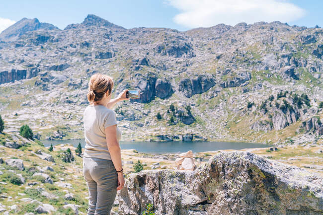 Back view anonymous female backpacker taking pictures on smartphone of amazing stony highlands with lake in Ruda Valley in Catalan Pyrenees — Fotografia de Stock