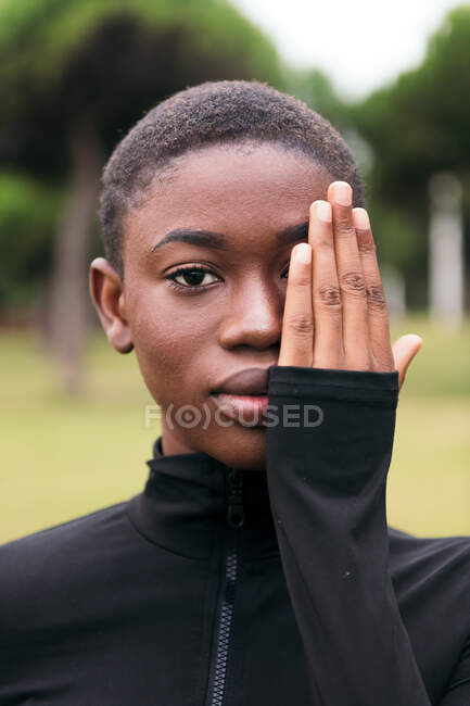 Young tender African American female with short hair covering face while looking at camera in town on summer day — Stock Photo