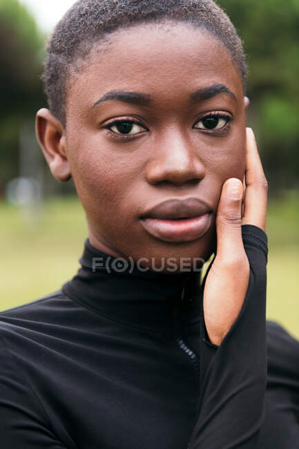 Jovem macio étnico feminino no preto vestuário com cabelo curto olhando para câmera no gramado urbano no verão — Fotografia de Stock