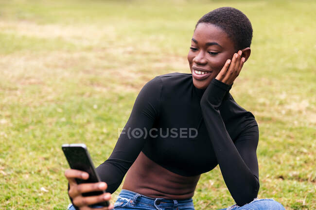 Молодая этническая женщина в повседневной одежде с беспроводной гарнитурой делает автопортрет на мобильном телефоне в парке — стоковое фото