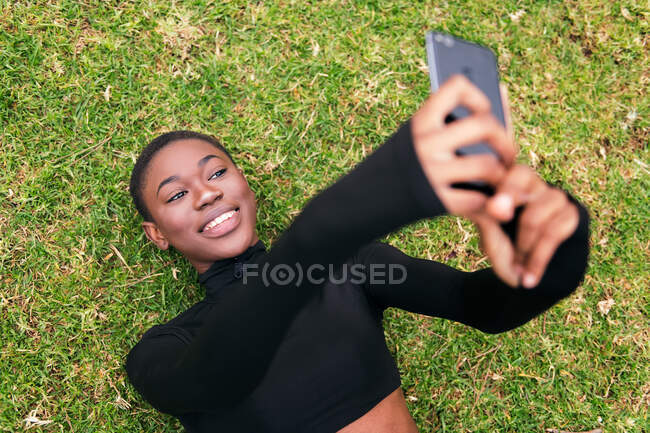 Jovem fêmea étnica em vestuário casual com fone de ouvido sem fio tirando auto-retrato no celular deitado em um parque — Fotografia de Stock