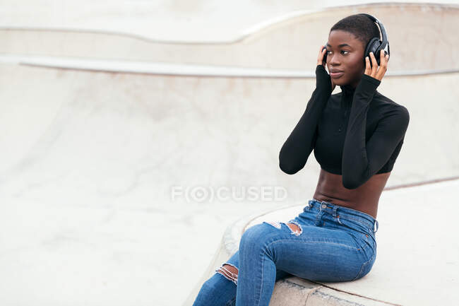 Vista lateral de una joven afroamericana contemplativa en jeans rasgados escuchando música de auriculares inalámbricos mientras mira hacia adelante - foto de stock