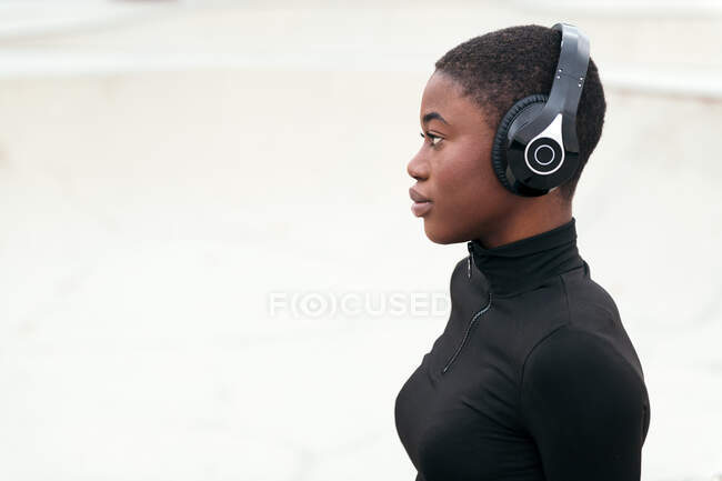 Вид сбоку на молодую созерцательную афроамериканку в разорванных джинсах, слушающую музыку из беспроводных наушников, глядя вперед — стоковое фото