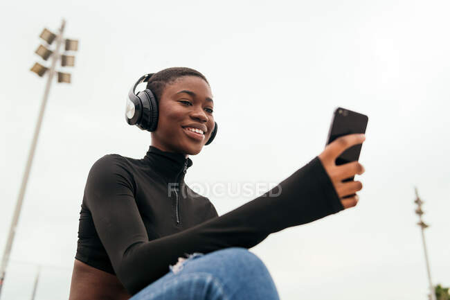 Low angle of young glad ethnic female in wireless headphones surfing internet on cellphone while listening to song in city — Stock Photo