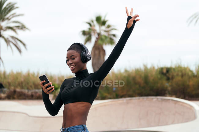 Low angle of young glad ethnic female in wireless headphones surfing internet on cellphone while listening to song in city — Stock Photo