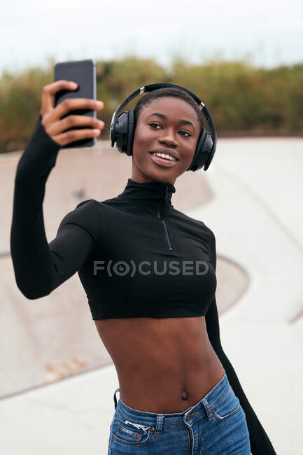 Молода етнічна жінка в повсякденному одязі з бездротовим гарнітуром взявши автопортрет на мобільний телефон в парку — стокове фото