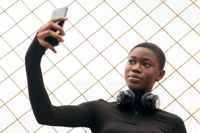 Junge Frau in lässiger Kleidung mit drahtlosem Headset porträtiert sich mit Handy gegen Gitterzaun — Stockfoto