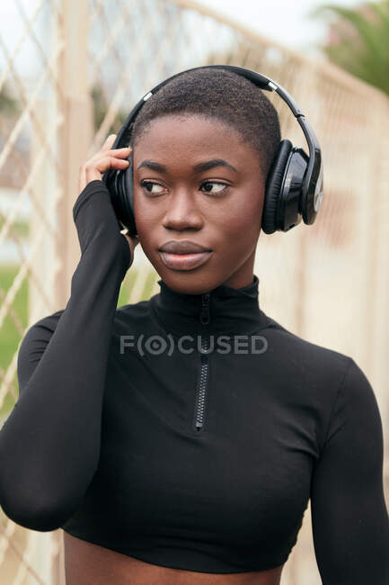 Cortar jovem sonhador preto fêmea ouvir música a partir de fones de ouvido sem fio enquanto olha para longe à luz do dia — Fotografia de Stock