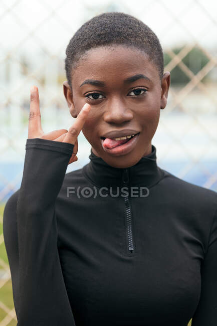 Smiling young African American female with stretched arm demonstrating fuck gesture in town on summer day - foto de stock