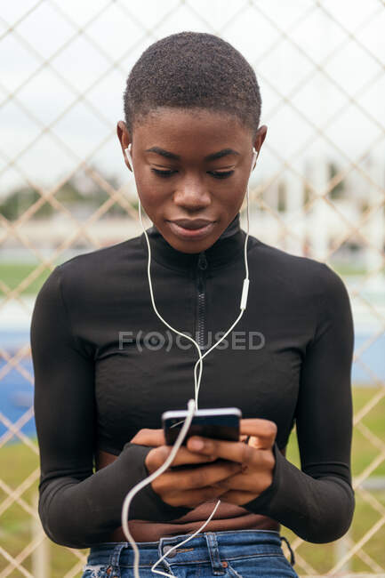 Young focused ethnic female text messaging on cellphone while listening to music against grid fence in city — Stock Photo