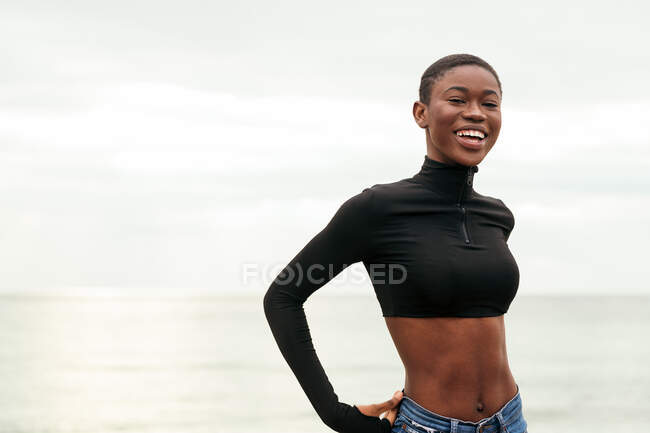 Jeune femme afro-américaine douce en vêtements décontractés regardant la caméra sur la côte de l'océan — Photo de stock