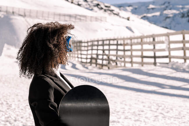 Happy ethnic sportswoman in stylish wear and protective glasses carrying snowboard on snowy mount in sunlight looking away — Stock Photo