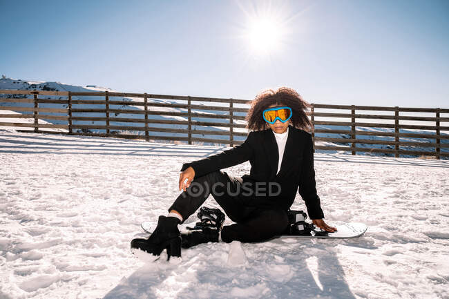 African American female athlete in trendy wear and protective glasses sitting on snowboard while looking at camera in winter — Stock Photo