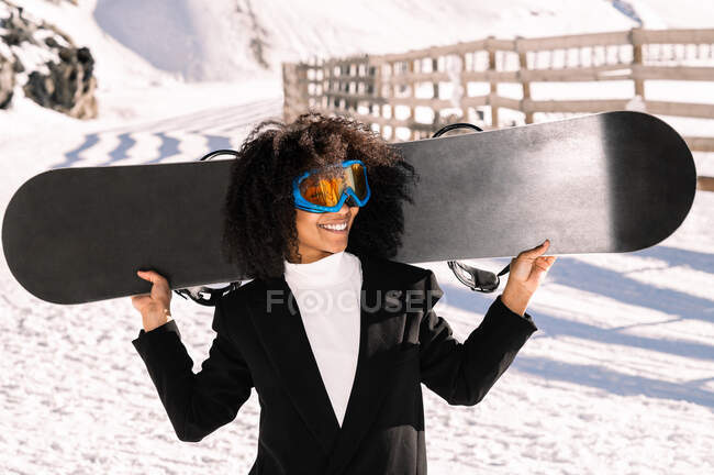 Happy ethnic sportswoman in stylish wear and protective glasses carrying snowboard on snowy mount in sunlight — Stock Photo