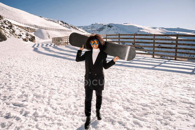 Happy ethnic sportswoman in stylish wear and protective glasses carrying snowboard on snowy mount in sunlight — Stock Photo