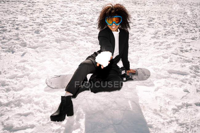 Cheerful African American female athlete in trendy wear and protective glasses sitting on snowboard while looking at camera in winter — Stock Photo